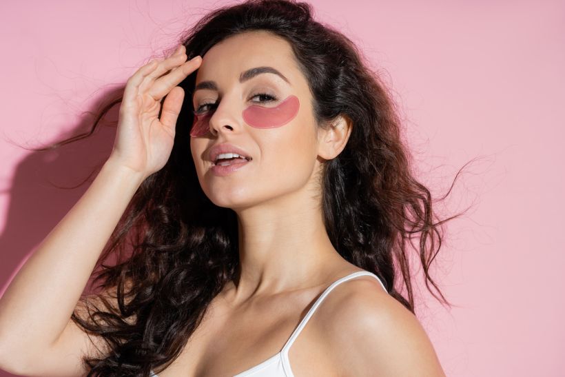 Pretty woman with hydrogel eye patches touching forehead on pink background / envato